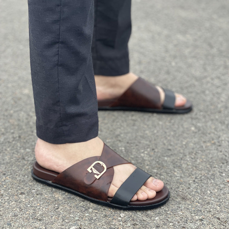 Men's brown leather sandals with a gold buckle detail and black strap worn with trousers.