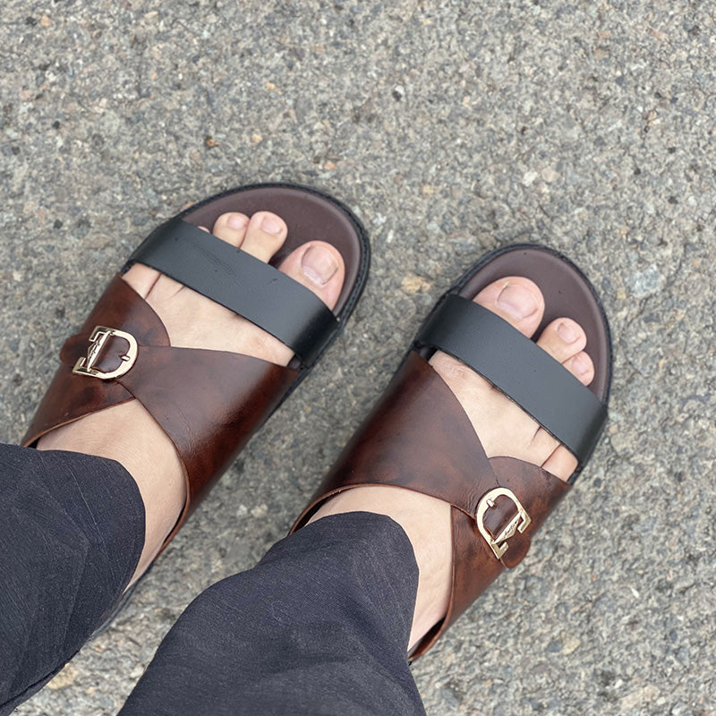 Handmade brown and black leather men's sandals with gold buckle detail on grey pavement.
