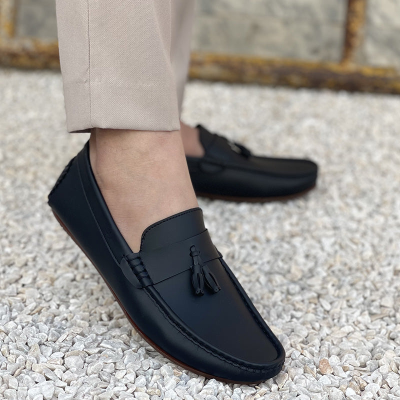 Man wearing black handmade leather tassel loafers with beige trousers on a pebble surface