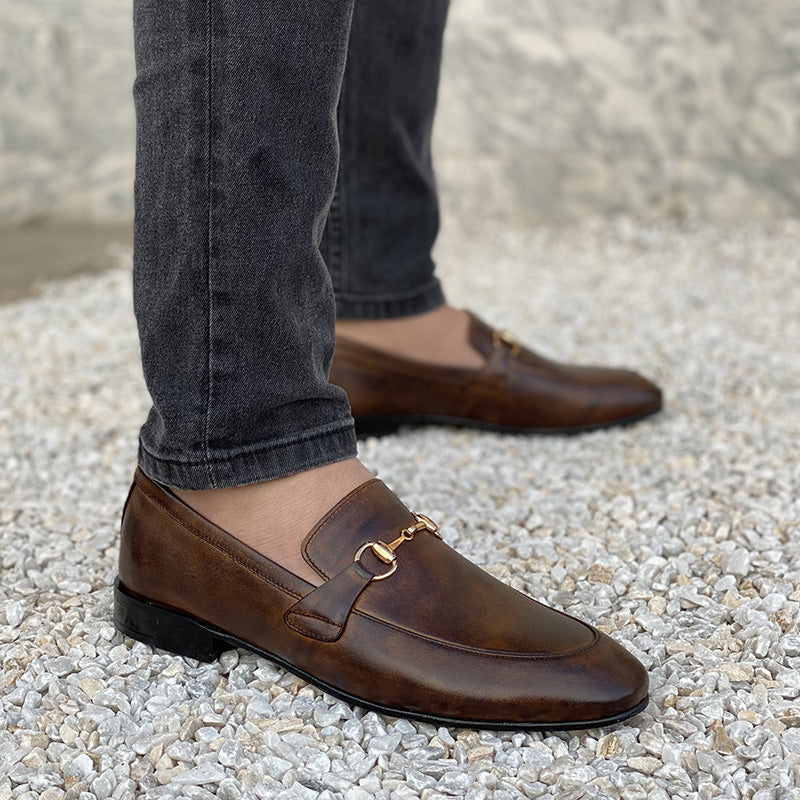Man wearing brown leather horsebit loafers with dark grey jeans on a white gravel surface