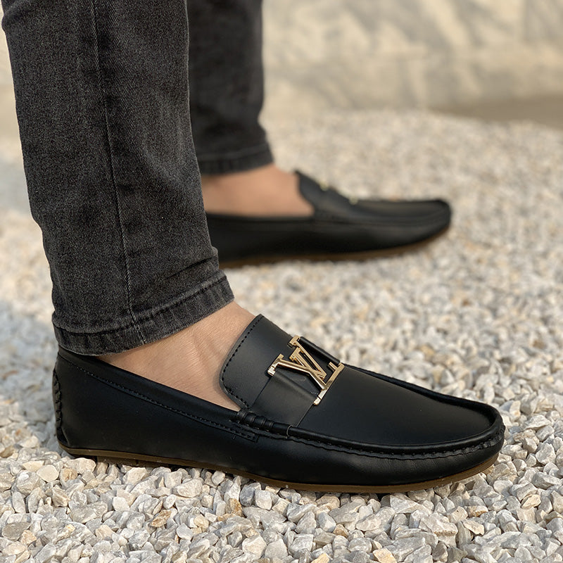 Man wearing black leather loafers with a gold metal buckle on a pebble surface