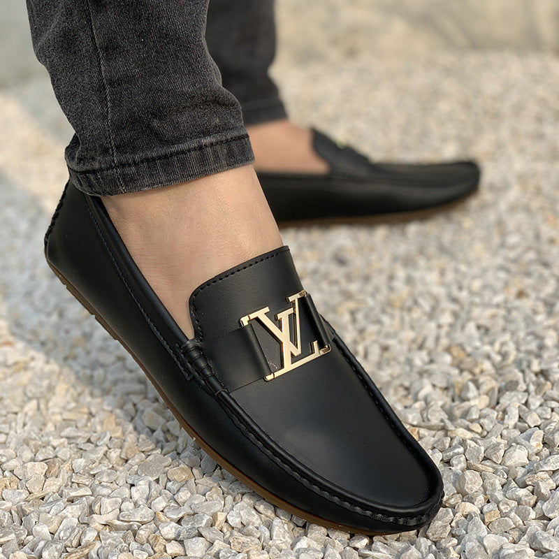 Close-up of black leather men's loafers with a gold logo buckle on a gravel background