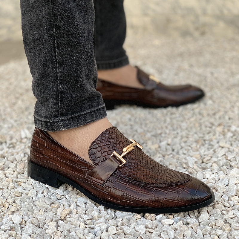 Men's brown croc-embossed leather loafers with gold buckle detail on white gravel.