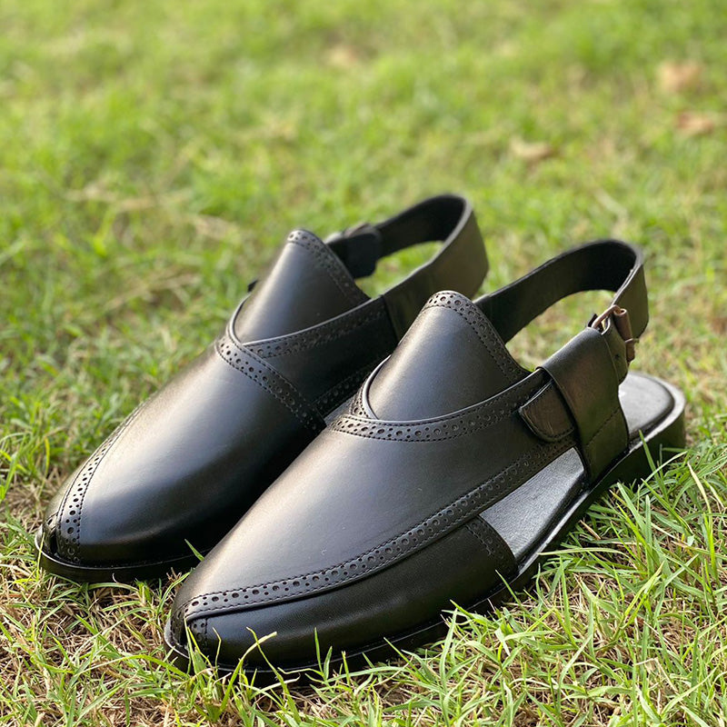 Handmade black leather men's mules with decorative brogue detailing on green grass.