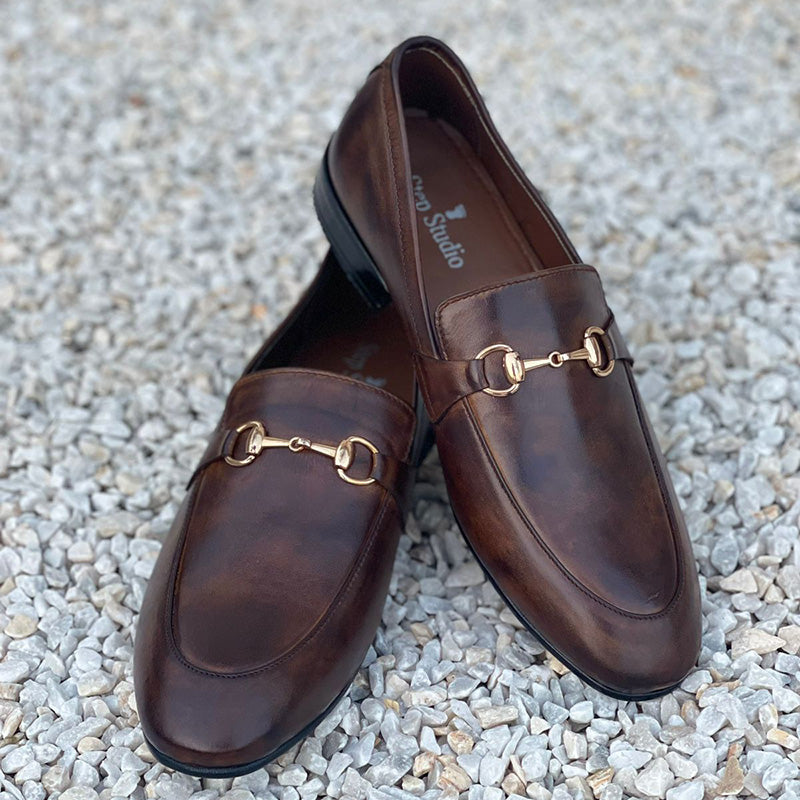 Pair of brown handmade leather loafers with gold horsebit hardware on a gravel surface
