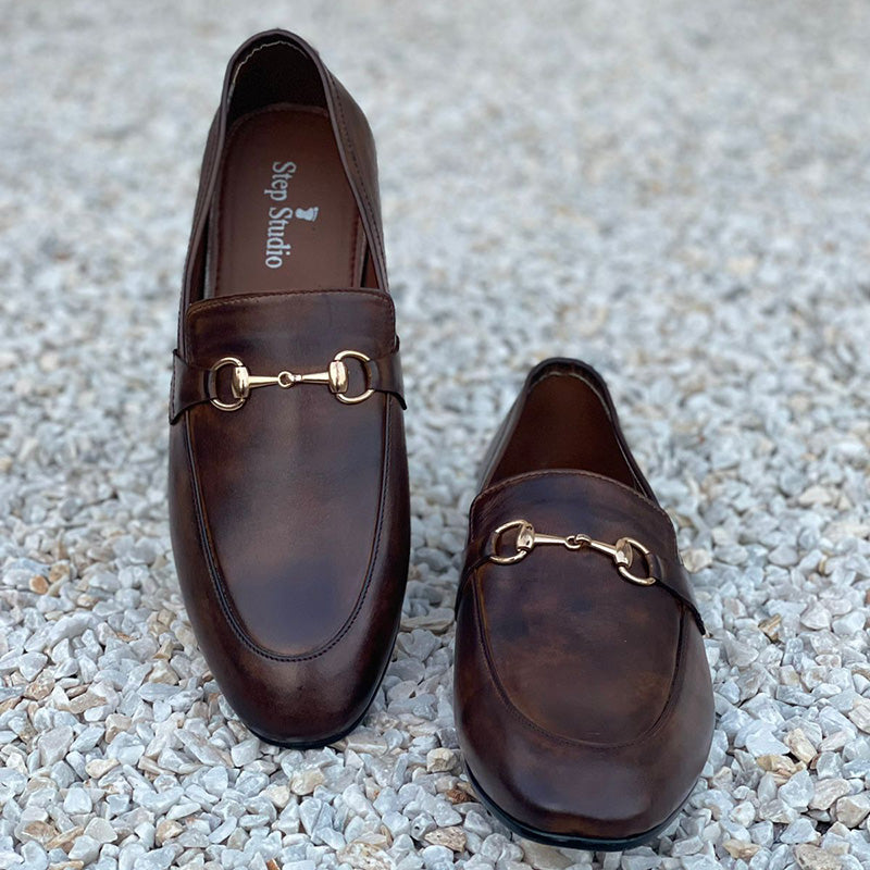 Pair of brown leather handmade men's loafers with gold horsebit detail on a gravel background