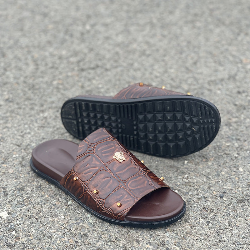 Brown crocodile texture leather men's slides with gold stud detailing on a grey asphalt background