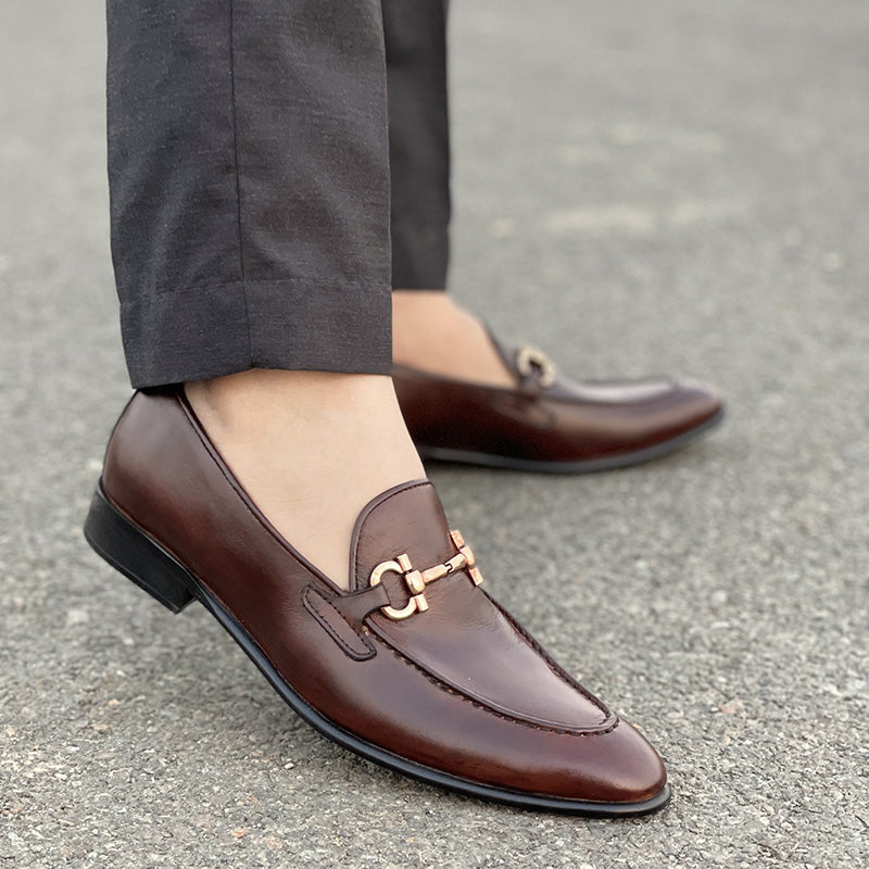Close-up of men's brown leather loafers with gold horsebit detail worn with grey trousers