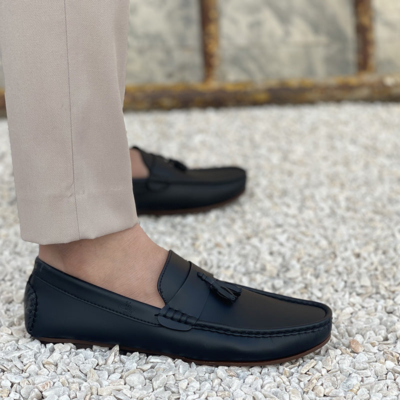 Close-up of a man wearing handmade black leather tassel loafers on a white pebble surface.