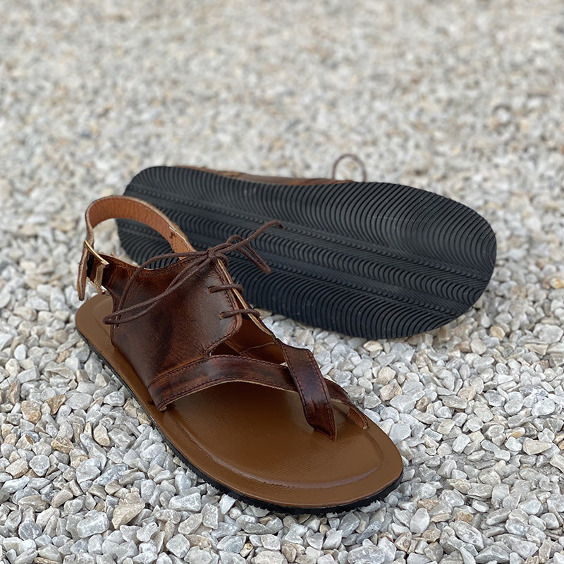 Handmade brown leather men's sandals with straps and black sole on a gravel background.