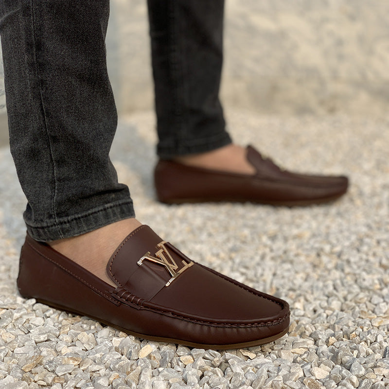 Man wearing brown leather loafers with a gold LV logo on a gravel surface