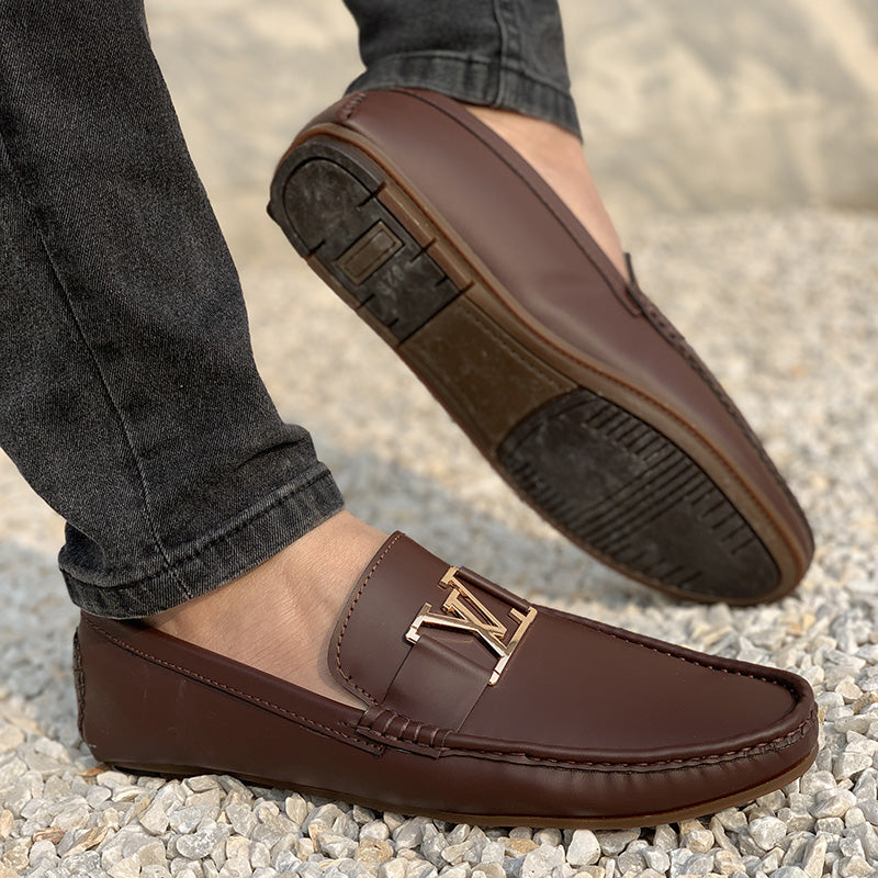 Close-up of a man wearing brown leather loafers with a gold buckle on a gravel surface.