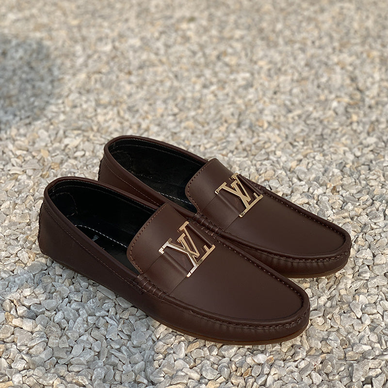 Pair of brown leather men's loafers with gold metallic hardware on a gravel background.