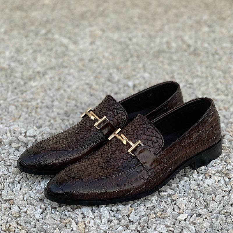 Pair of brown crocodile leather loafers with gold metal hardware on a gravel background