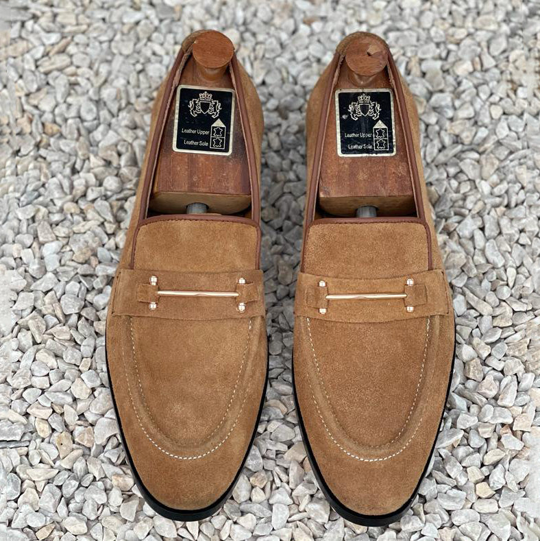 Handmade tan suede men's loafers with metal bit detailing on a pebble background